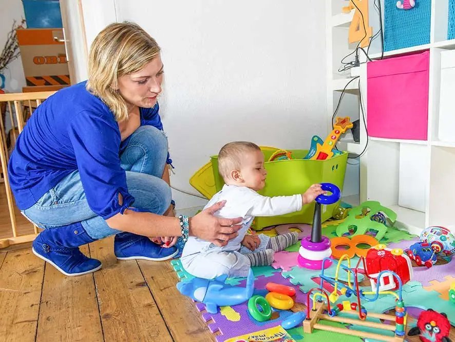 Eine Frau sitzt mit einem Baby in einer bunten Spielecke. Das Baby schiebt einen blauen Ring über ein Kegelförmiges Spielzeug.