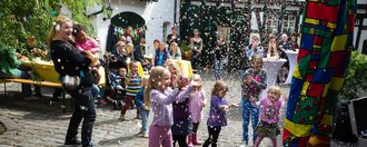 Bei einer Clownvorstellung im Freien jubeln Kleinkinder in einem Konfettiregegen. Im Hintergrund stehen Erwachsene vor einem Fachwerkhaus und beobachten die Szene.