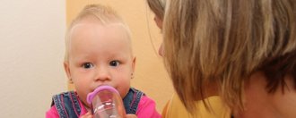 Eine Frau hält ein Baby auf dem Arm, welches gerade an einer Trinkflasche nuckelt. Die Frau blickt das Baby an.