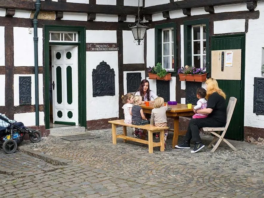 Zwei Frauen sitzen mit vier Kleinkindern vor einem alten Fachwerkhaus um einen Tisch und unterhalten sich